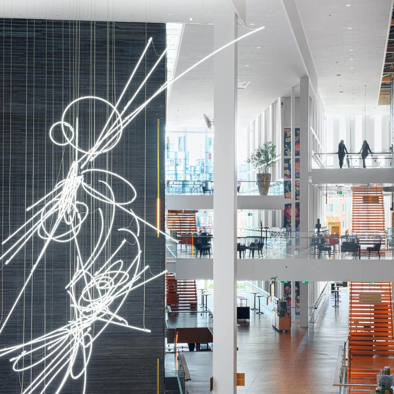 Futuristisk lysskulptur henger i et romslig atrium med moderne, flerprangende gangveier og sitteplasser. Store teksturerte vegger gir en skarp bakgrunn til det lyse, åpne rommet.