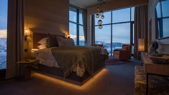 A bed with decorative cushions sits in a warmly lit bedroom, surrounded by large windows showing snowy mountains. A seating area and wooden furniture complete the cozy, elegant environment.
