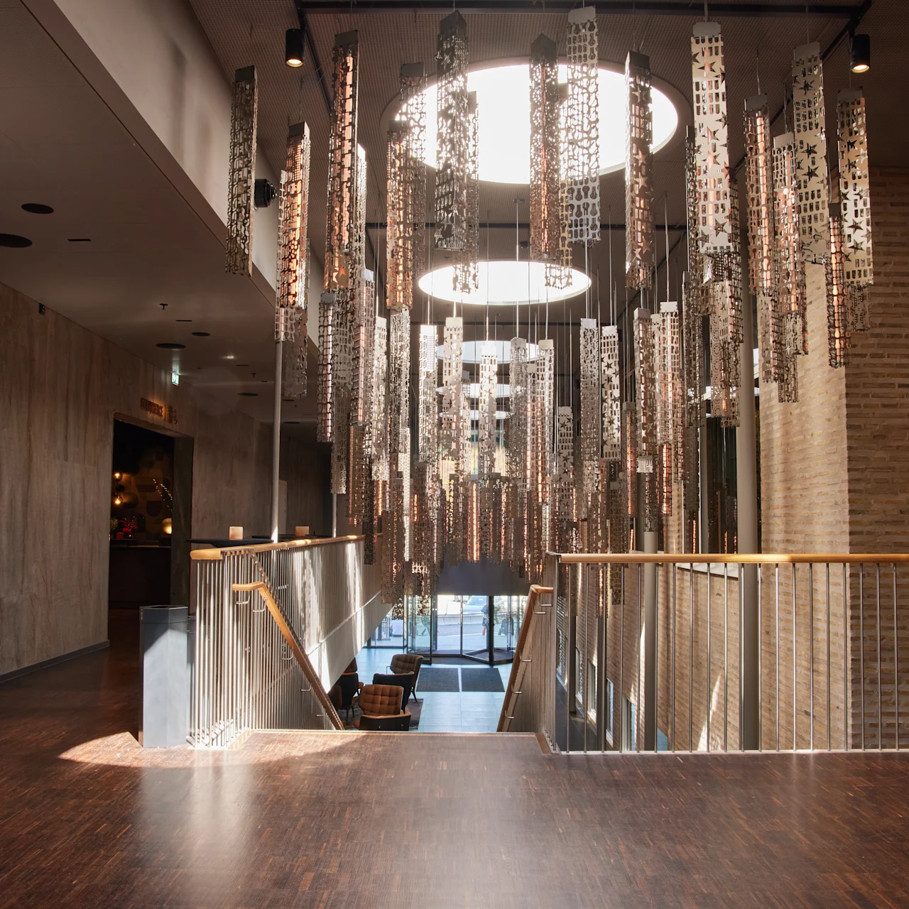 Ett hängande ljusinstallation svävar ovanför en trappa med metalräcken. Det moderna interiören har trägolv och tegelväggar, belysta av naturligt ljus från runda takfönster.