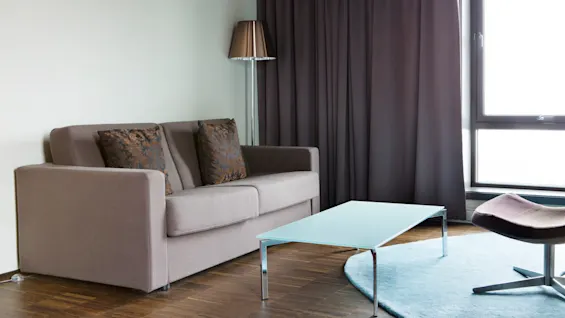 En grå sofa med to mønstrede puter står ved siden av en stålampe. I nærheten hviler et glassbord på et blått teppe i en moderne stue.