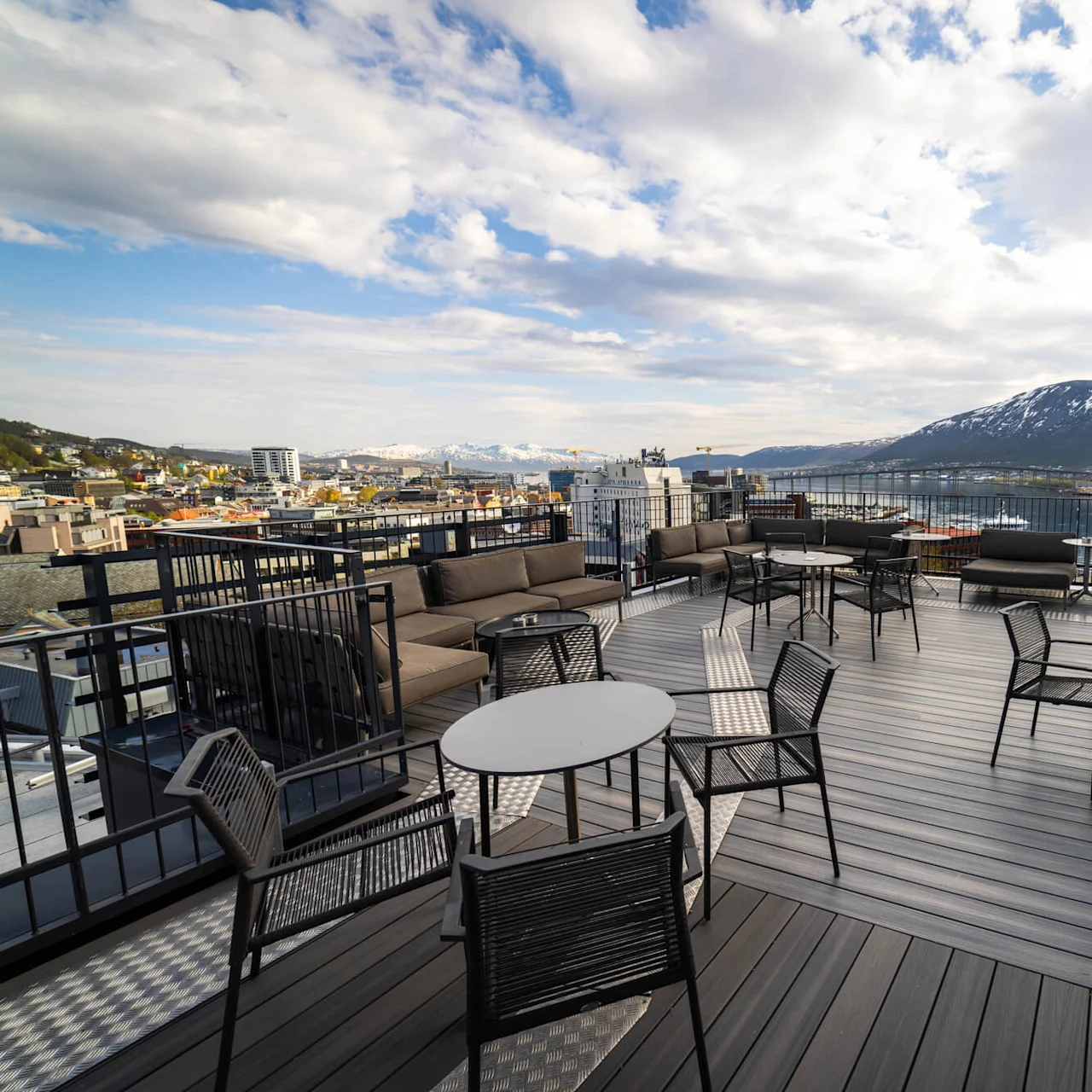 Udendørs terrasse med tomme borde og stole, omgivet af glasruder, med udsigt over en by med farverige bygninger, en vandmasse og sneklædte bjerge under en delvist skyet himmel.