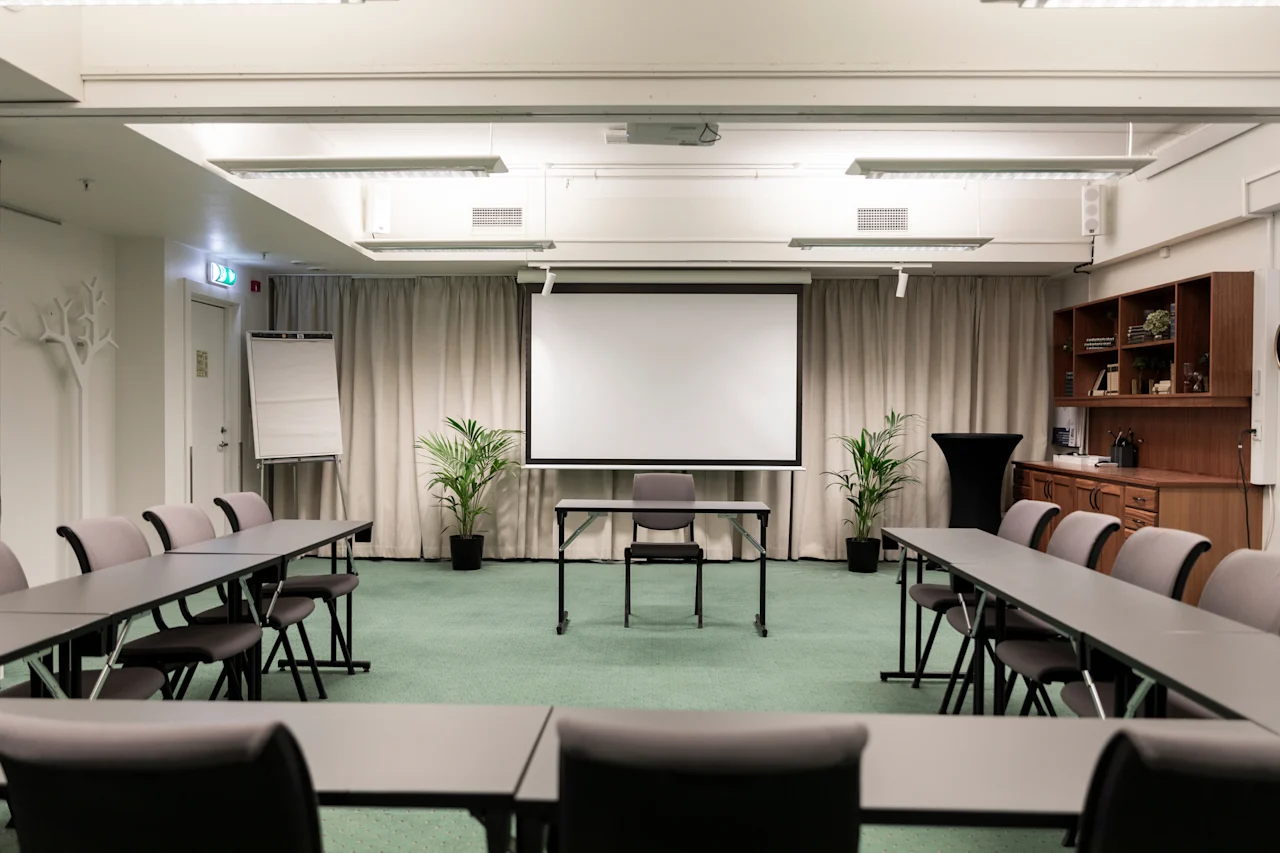 A spacious conference room at Home Hotel Grand Bodø with tables and chairs arranged for a meeting, facing a large projector screen.
