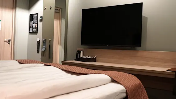A neatly made bed, covered with white linens and a brown throw blanket, faces a wall-mounted, large flat-screen TV in a modern, brightly lit hotel room.
