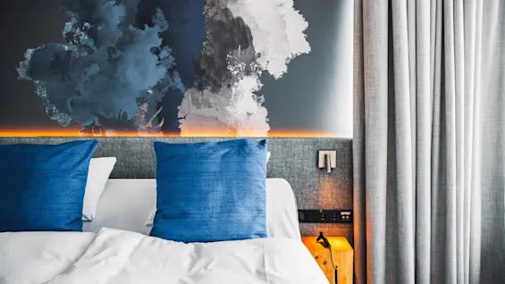 A bed with blue pillows is beneath a large abstract mural. Soft lighting highlights a gray headboard. Curtains frame one side of the room.