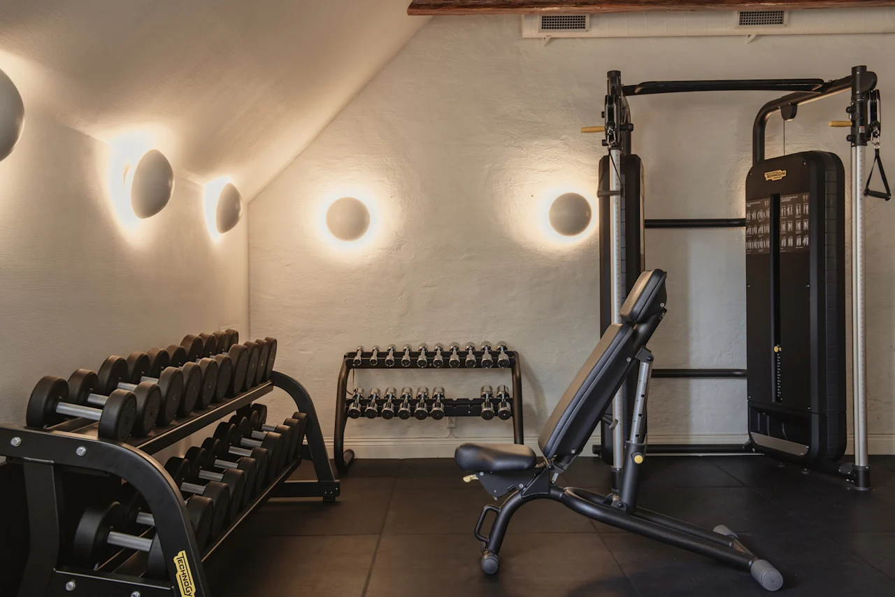 Ett väl upplyst gym med olika träningsutrustning, inklusive hantelställ, en träningsbänk och en multifunktionell träningsmaskin.