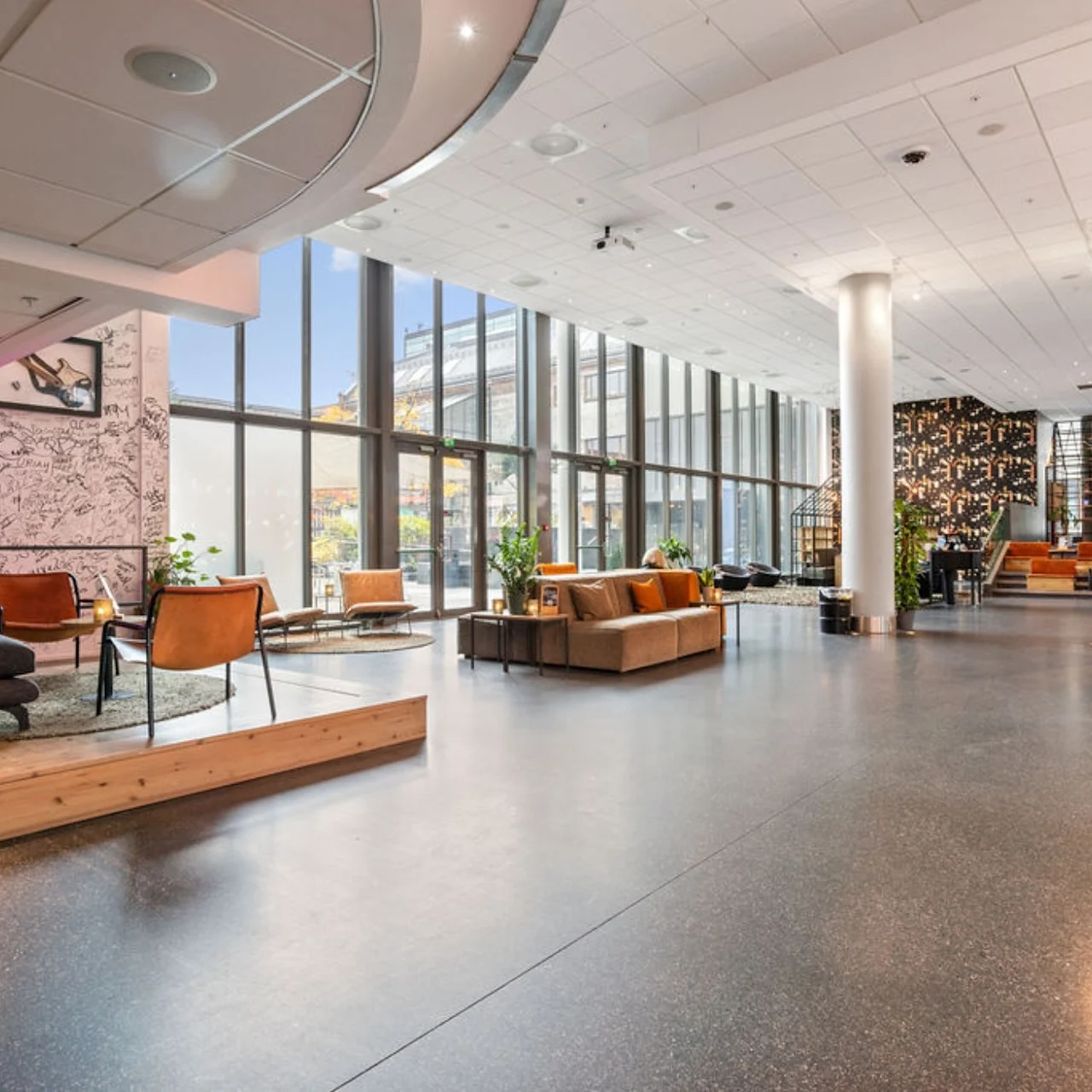 A spacious, modern lobby features stylish seating arrangements and large floor-to-ceiling windows providing natural light. Plants and contemporary decor enhance the aesthetic, creating a welcoming atmosphere.