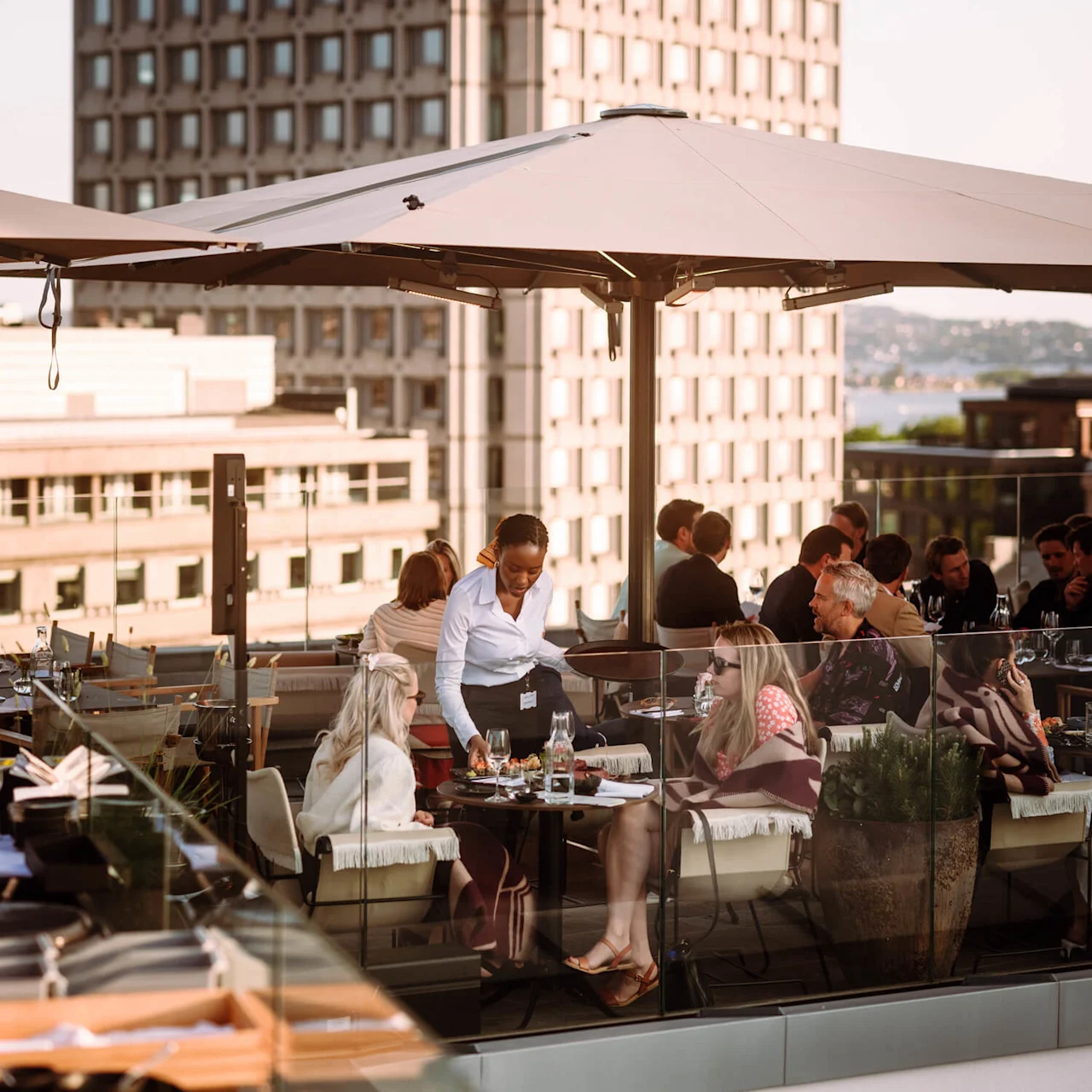 En tjenerinde betjener gæster på en tagterrasse med bysilhuet i baggrunden.