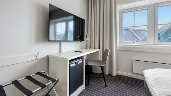 A modern hotel room with a wall-mounted TV, a desk with a chair, and a glimpse of a bed; the window reveals a sunny day.