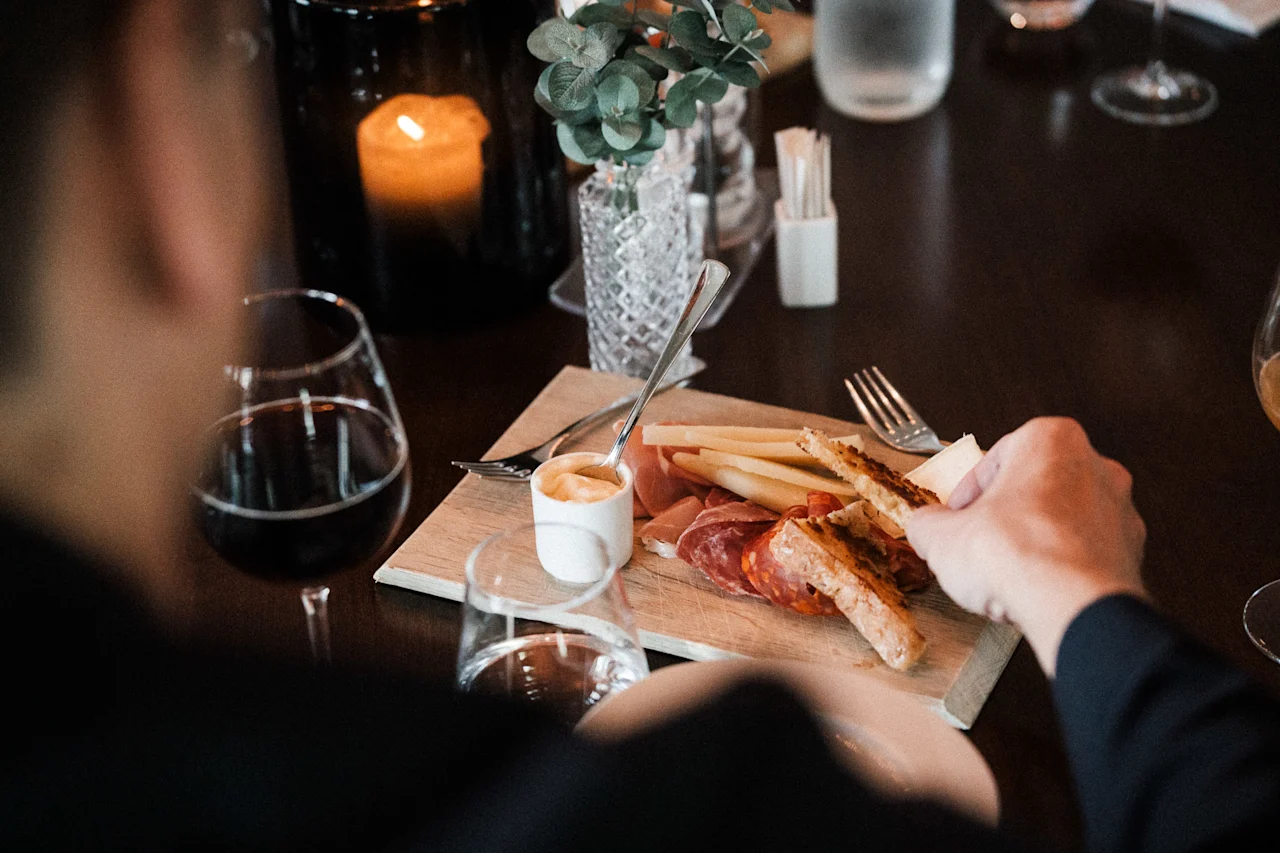 En person nyter et forrettsfat med spekemat, ost og brød, akkompagnert av vin i en koselig, levende lyssetting.
