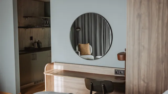 En rund spegel ovanför ett skrivbord i trä speglar en fåtölj med en kudde. Miljön är ett modernt, minimalistiskt rum med ett pentry synligt till vänster.