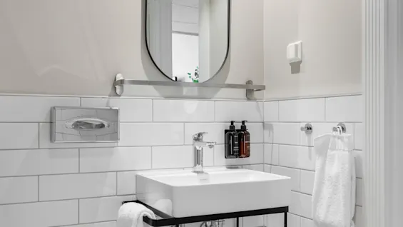 A clean and modern bathroom featuring a white sink, an oval mirror, and subway tiles, with soap and fresh towels.