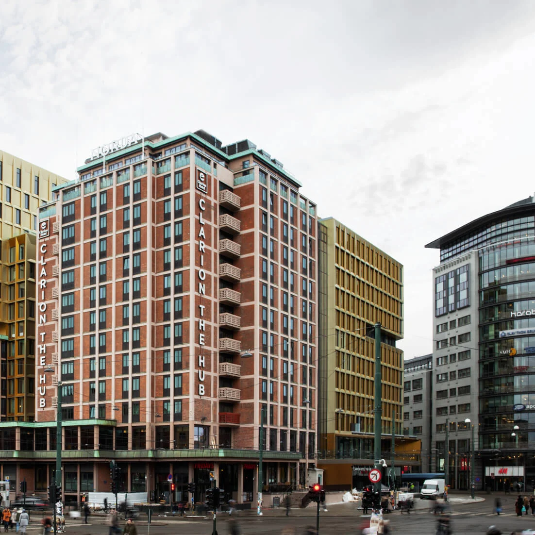 Clarion The Hub building stands tall, displaying "CLARION THE HUB" on its facade in an urban setting. Surrounding it are modern and rounded office buildings. People and vehicles are visible on the street. 