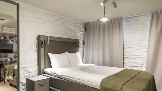 Viihtyisä hotellihuone, jossa on mukava sänky, valkoiset vuodevaatteet ja vihreä peitto, sekä tekstuuriseinät ja pehmeä valaistus.