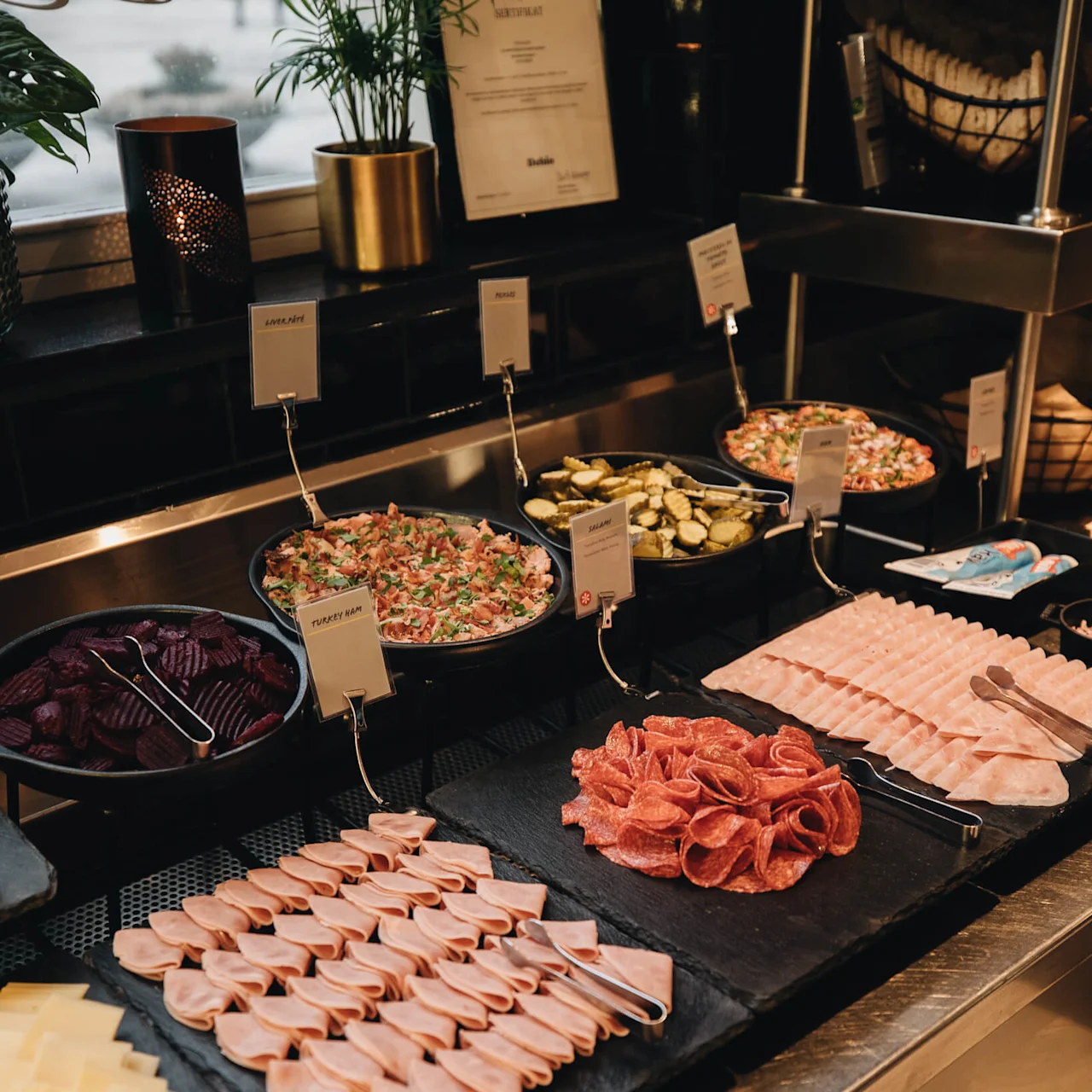 A deli counter displays sliced meats, cheeses, and salads with labels like "Turkey Ham" and "Sallad" in a restaurant setting with plants and metal containers.