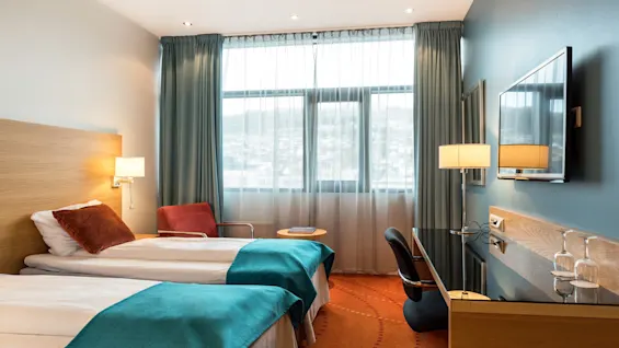 To enkeltsenger med turkise tepper står i et lyst hotellrom, med en koselig stol, skrivebord, flatskjerm-TV og stort vindu med lette gardiner.