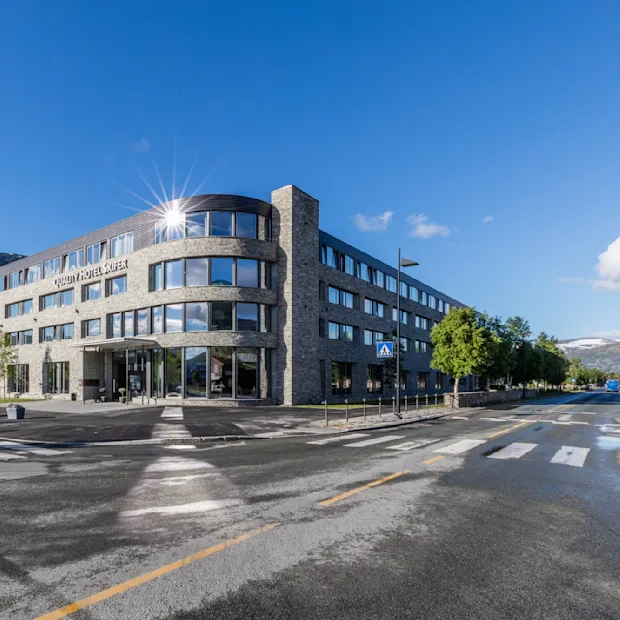 En moderne fleretagers bygning med navnet "Scandic Valdres Hotel" står på et gadehjørne, med reflekterende vinduer omgivet af træer, bjerge og en klar blå himmel.