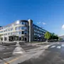 En moderne fleretagers bygning med navnet "Scandic Valdres Hotel" står på et gadehjørne, med reflekterende vinduer omgivet af træer, bjerge og en klar blå himmel.