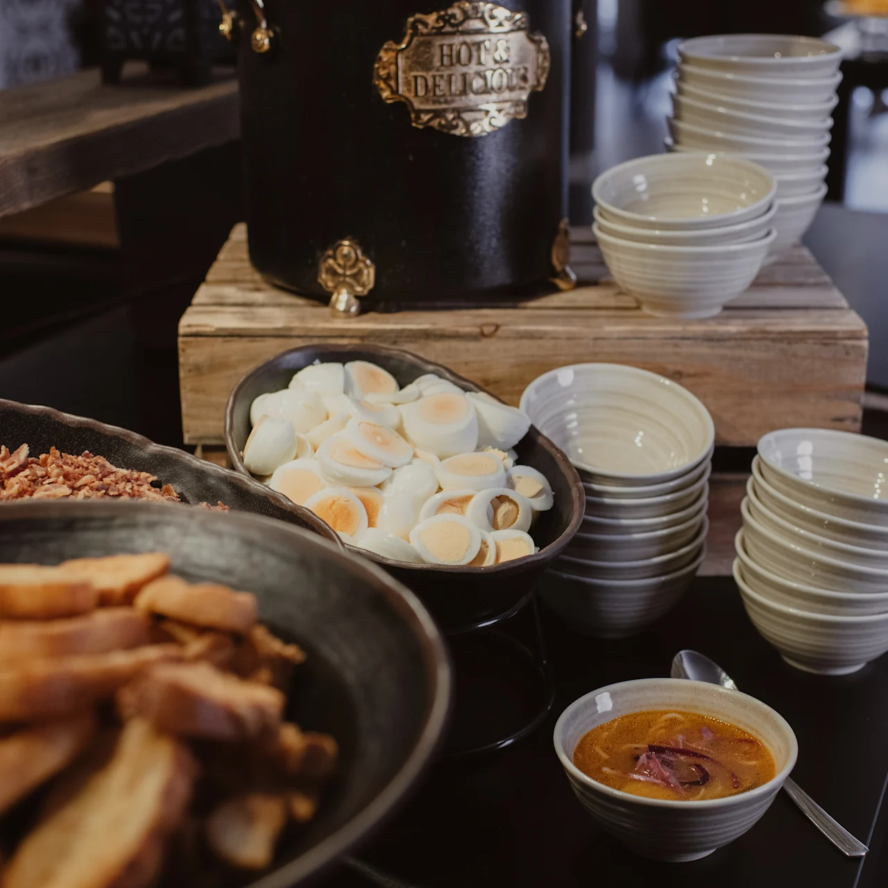A display of food includes sliced hard-boiled eggs, crispy bread, and bacon bits in bowls. Nearby, a pot labeled "Hotel Deluxe" stands on wood, surrounded by stacked bowls.
