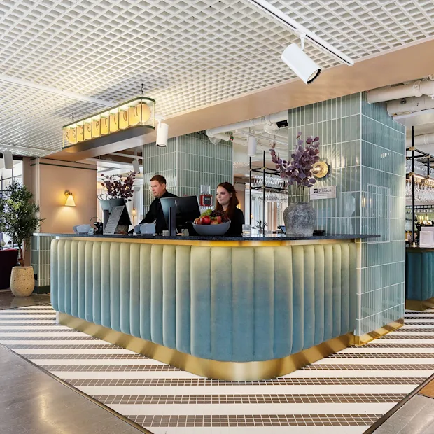 To ansatte som står bak en moderne hotellresepsjon og ønsker gjester velkommen.