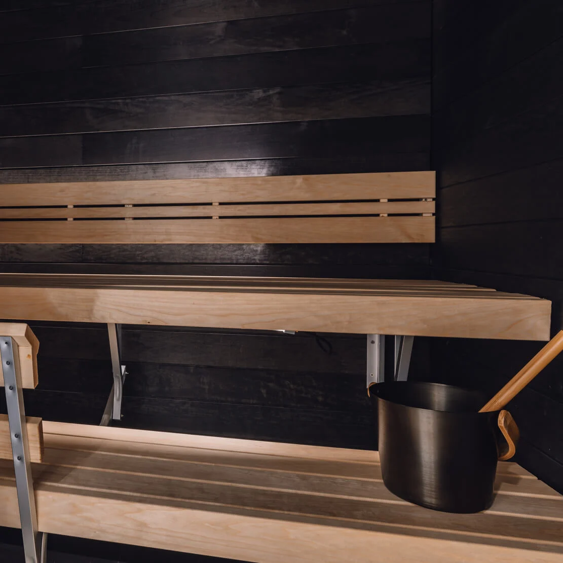 Wooden benches sit quietly in a dimly lit sauna, with a black bucket and ladle placed on the lower bench, surrounded by dark wooden walls.
