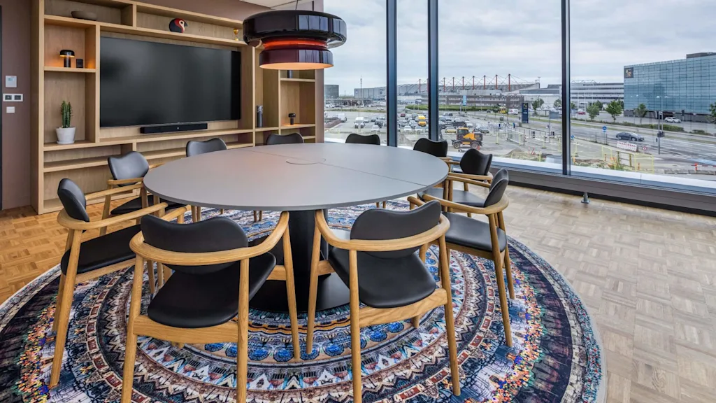 Ett modernt konferensrum med ett stort bord, stolar och en bred TV-skärm, med utsikt över ett urbant landskap genom golv-till-tak-fönster.