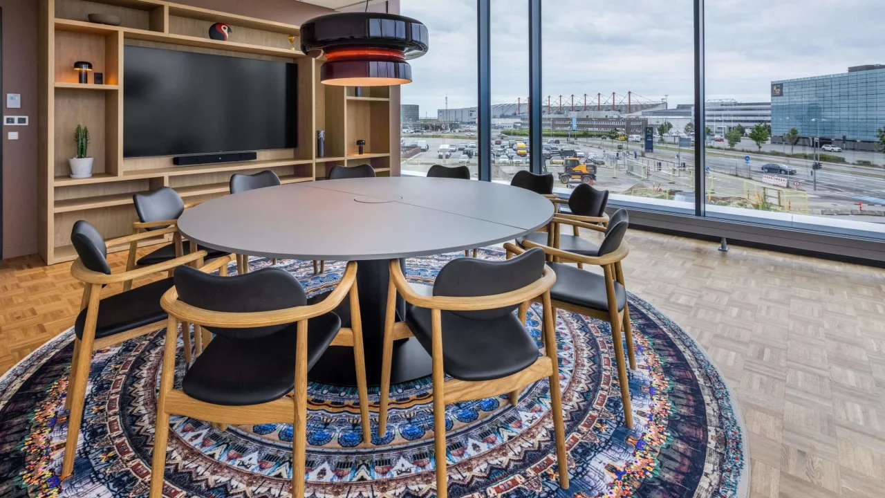 A modern conference room with a large table, chairs, and a wide television screen, overlooking an urban landscape through floor-to-ceiling windows.