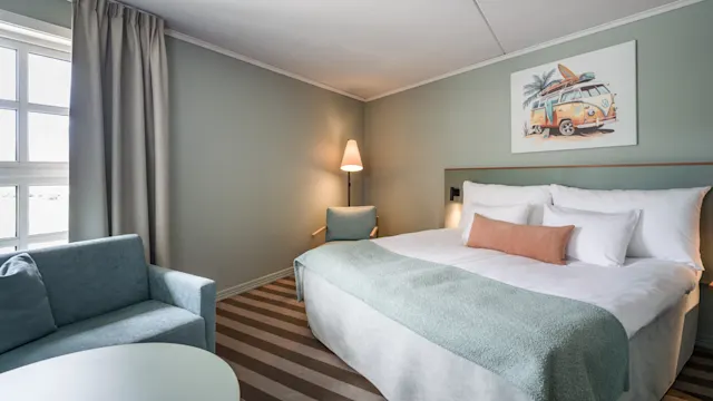 En pent redd seng med et lyseblått teppe og flere puter står ved siden av en lampe i et svakt opplyst rom med stripete teppe, et vindu og en sittegruppe.