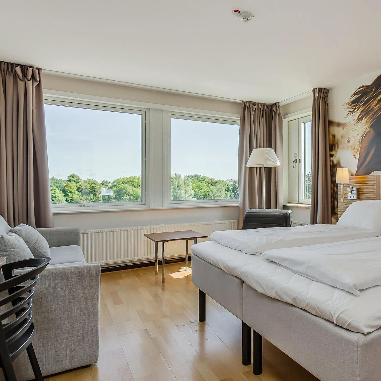Siististi pedattu parisänky valkoisilla liinavaatteilla sijaitsee hotellihuoneessa, jossa on suuret ikkunat, vihreä näkymä, puulattia, seinämaalaus ja yöpöydän lamput.