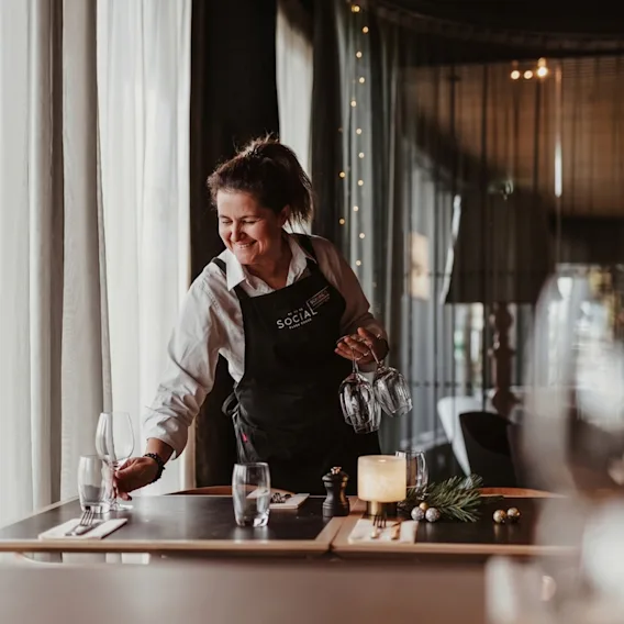 En smilende servitør dekker på til middag ved et koselig restaurantbord nær et vindu.