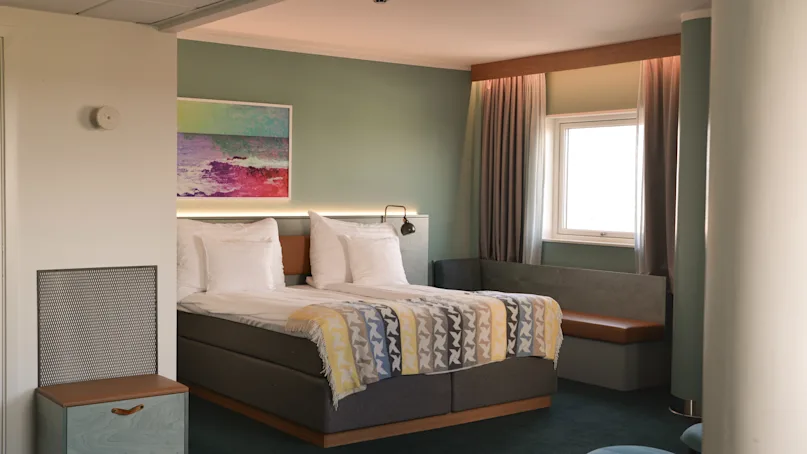 A neatly made double bed with patterned blankets stands in a stylish room featuring a green wall, framed art, and a small window seat bathed in soft natural light.
