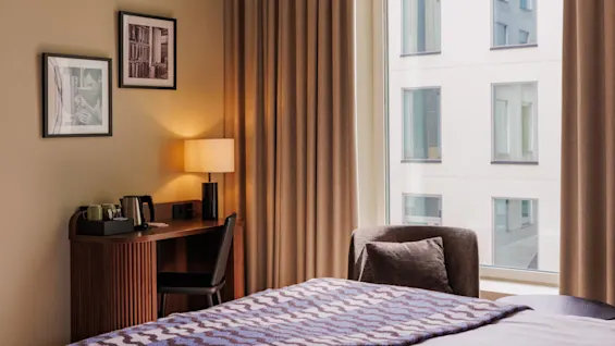 A neat hotel room with a bed, desk, and chair near a window with thick curtains and framed photos on the wall.