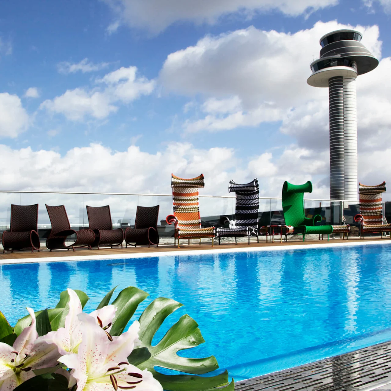 A rooftop swimming pool has blue water, surrounded by modern lounge chairs. A tall control tower is visible in the background under a partly cloudy sky.