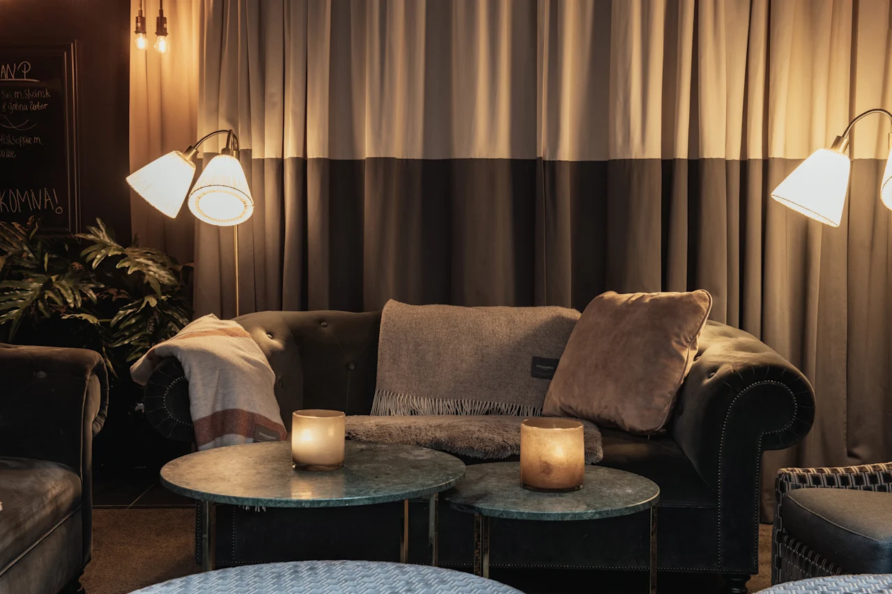 Et hyggeligt loungeområde med en mørk sofa, to runde borde med glødende stearinlys og varm belysning fra gulvlamper. KOMNA!