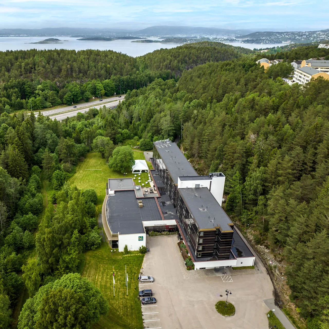 Aerial view of a multi-story building surrounded by dense green forest, with a road nearby and a body of water in the distance.