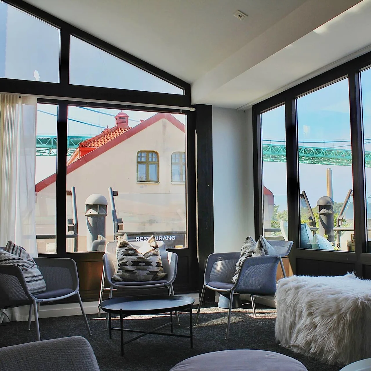 A cozy seating area with chairs and a fur-covered bench, surrounded by large windows overlooking a building with a red roof and a green bridge. Text: "RESTURANG".
