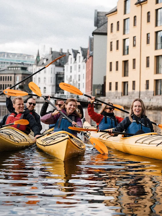 Folk i kajakker på en kanal, smilende og nyter aktiviteten med bygninger i bakgrunnen, inkludert et hotell.