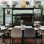 Stolar och bord är arrangerade i ett modernt, inomhus café med gröna väggar. Krukväxter och böcker dekorerar hyllor, medan naturligt ljus strömmar in genom stora fönster ovanför.