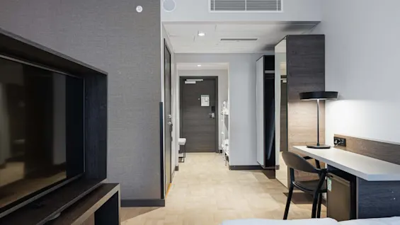 Ett hotellrum med en stor TV på ett stativ, ett skrivbord med lampa och stol, och en korridor som leder till en stängd dörr.