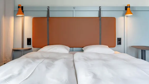 En pænt redt dobbeltseng med hvidt sengetøj er centreret under et brunt sengegærde. To orange lamper er monteret på begge sider og lyser det moderne, minimalistiske soveværelse op.