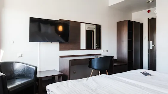 Ett modernt hotellrum har en stor väggmonterad TV ovanför ett skrivbord med stol, intill en säng och garderob.