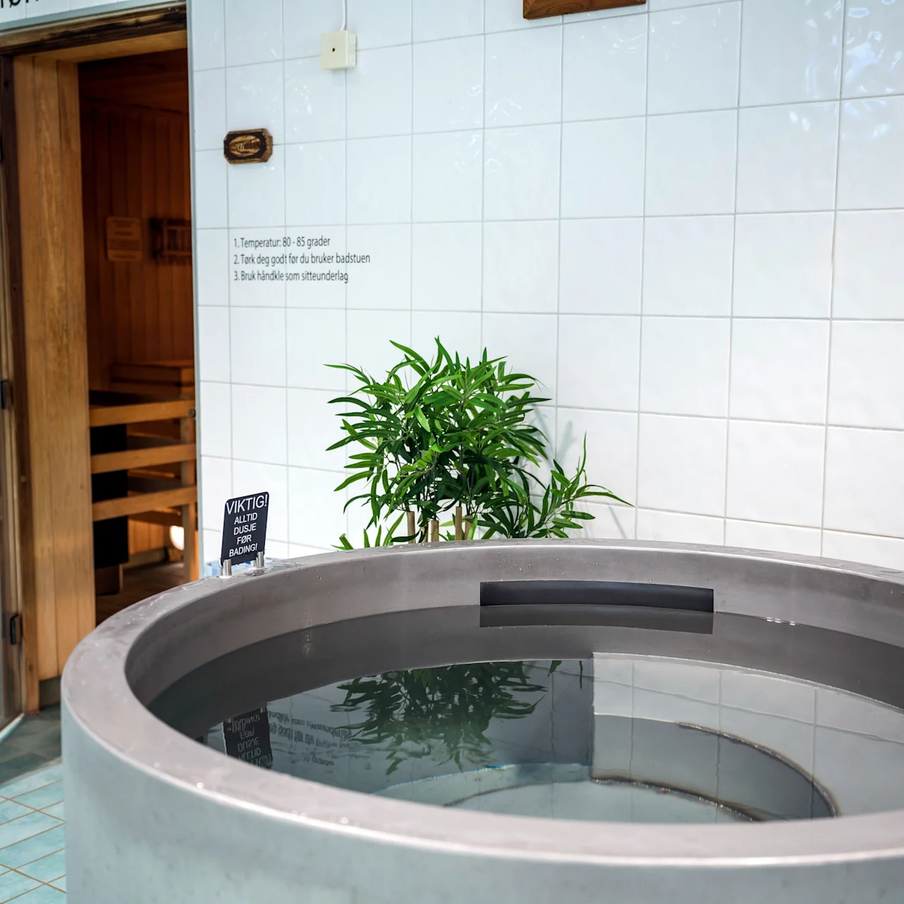 A round hot tub filled with water sits next to a sauna. White tiled walls surround the area. Text: "VIKTIG! ALLTID DUSJ FØR BADING."