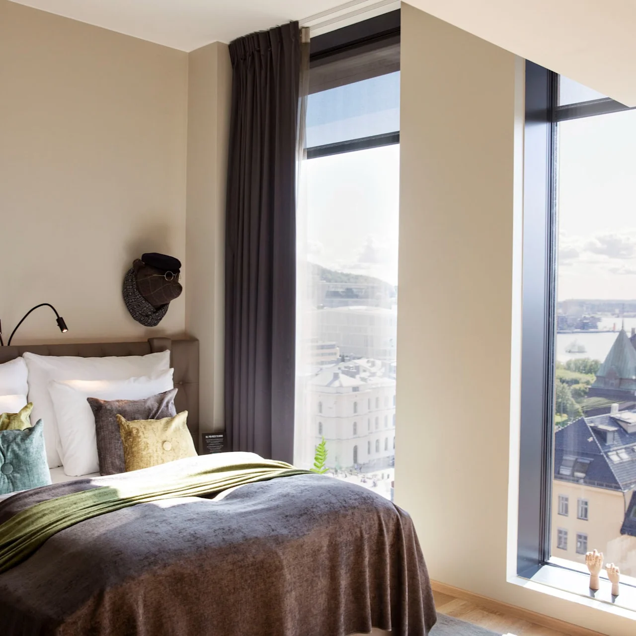 A neatly made bed with colorful pillows and a brown throw blanket stands by large windows. The windows offer a cityscape view of rooftops and a distant body of water under a clear sky.