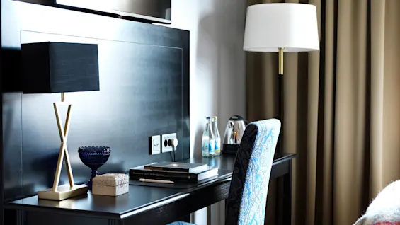 Et skrivebord med en lampe og bøker står ved siden av en blåmønstret stol, under en veggmontert TV; scenen er i et moderne rom med seng og gardiner.
