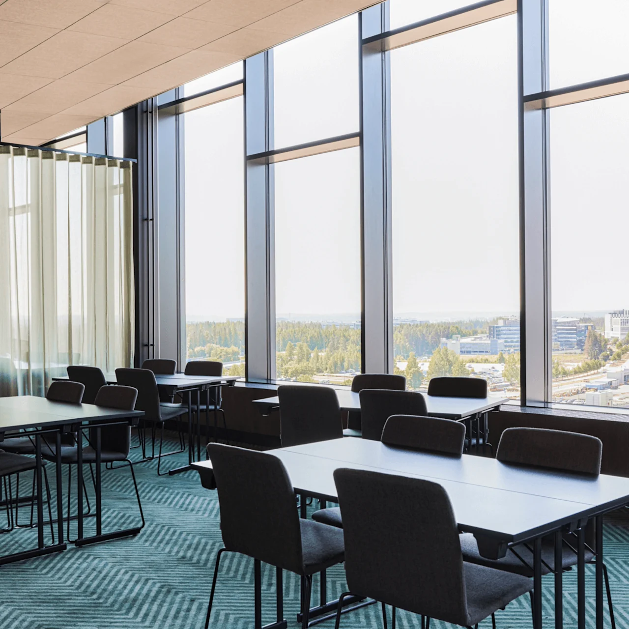 Meeting tables sit quietly in a spacious room with floor-to-ceiling windows and light curtains, overlooking an urban landscape with buildings in the distance.
