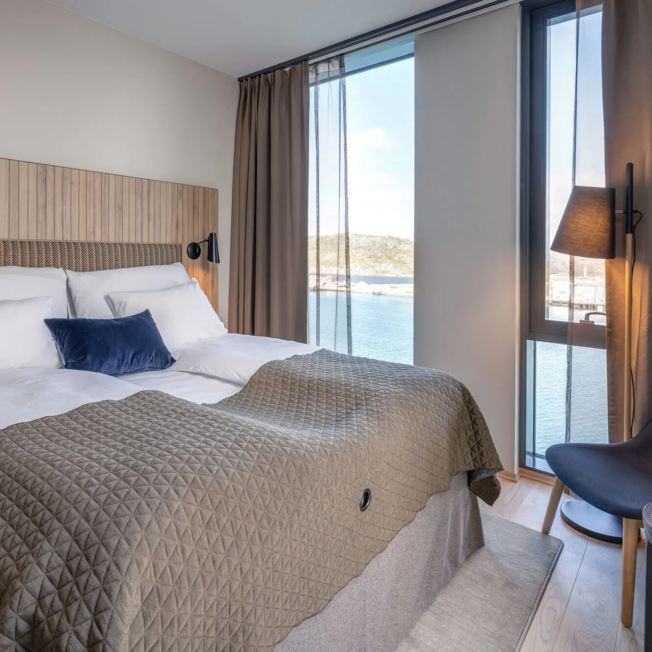 A neatly made bed with white linens and a grey quilt, positioned in a bright room with wooden accents, overlooks a calm body of water through large windows.