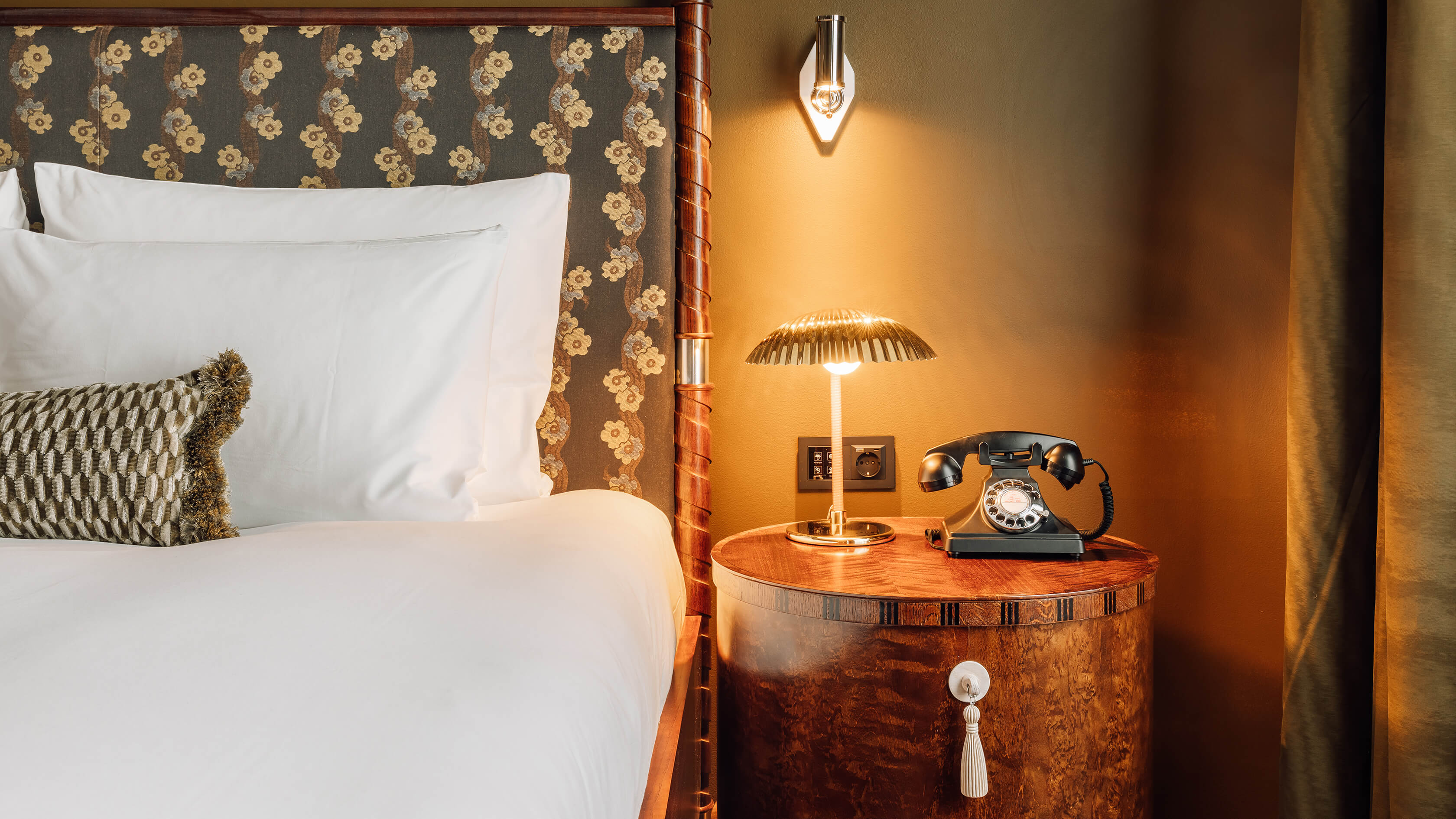 A neatly made bed with a vintage phone and lamp on a nightstand, in a cozy room with elegant decor.