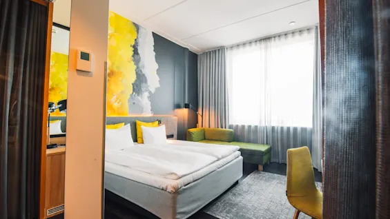 A bed with white linens stands in a modern hotel room featuring a colorful, abstract wall mural. Large window drapes allow natural light, while a green chair and sofa add seating.