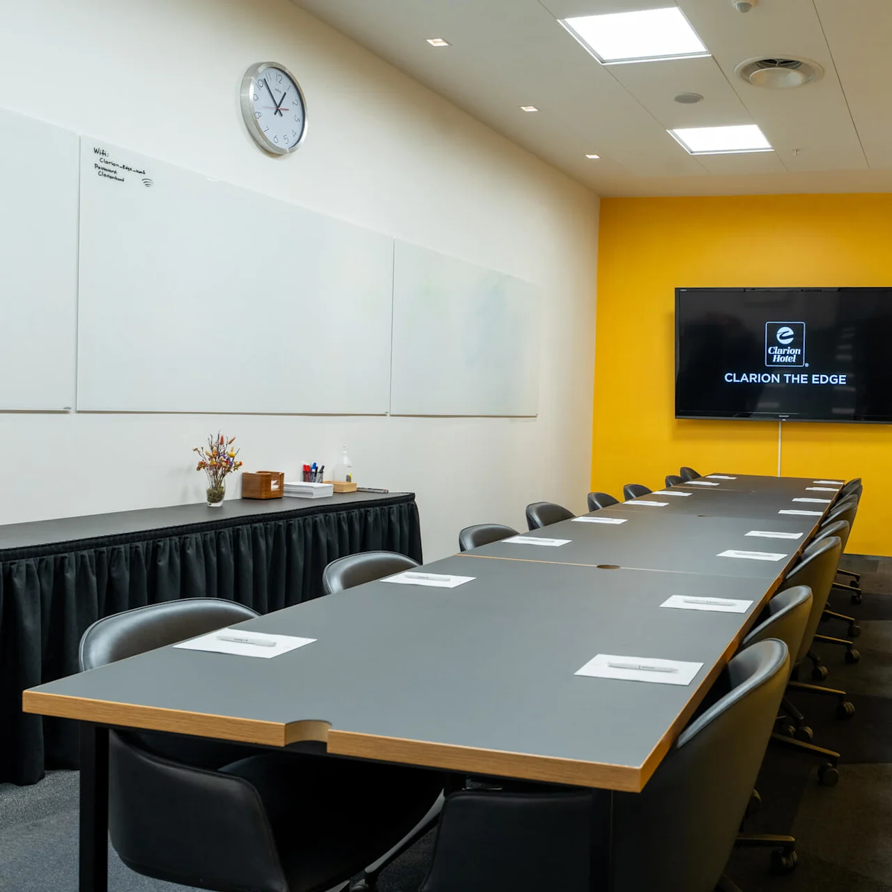 Konferencebord omgivet af stole, opstillet med navneskilte, i et gult malet rum. TV'en viser "Clarion Hotel CLARION THE EDGE." En whiteboard og ur på en anden væg.