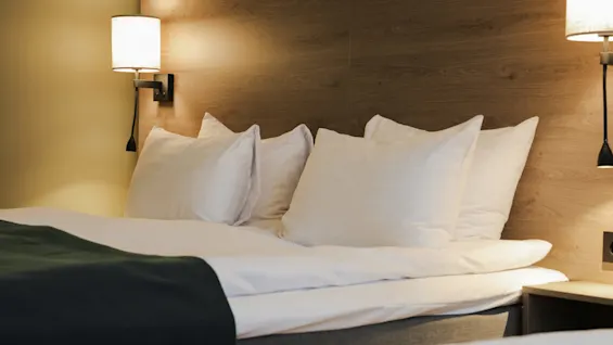 A neatly made bed with white pillows and a dark throw rests against a wooden headboard, flanked by two wall-mounted lamps illuminating the cozy room.