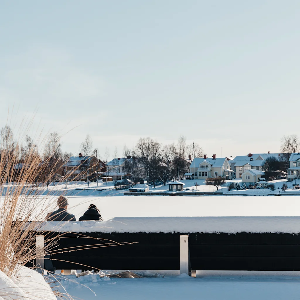 Kaksi ihmistä istuu lumisella penkillä ja katsoo jäätyneen järven yli talojen suuntaan kirkkaan talvitaivaan alla.